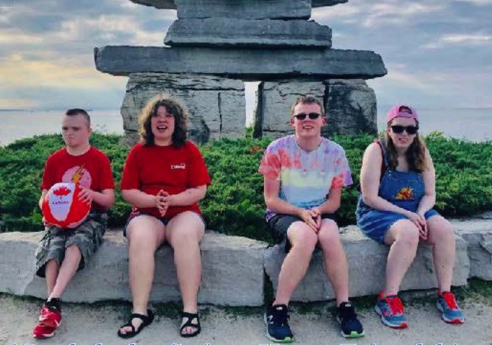 Group of four young adults sitting on a stone ledge in front of a large stone sculpture, smiling and enjoying time outdoors.